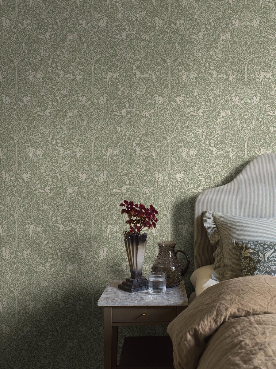 Under the Elder Tree wallpaper by Borastapeter, a sage green elder tree with forest animals behind a bed and a marble side table
