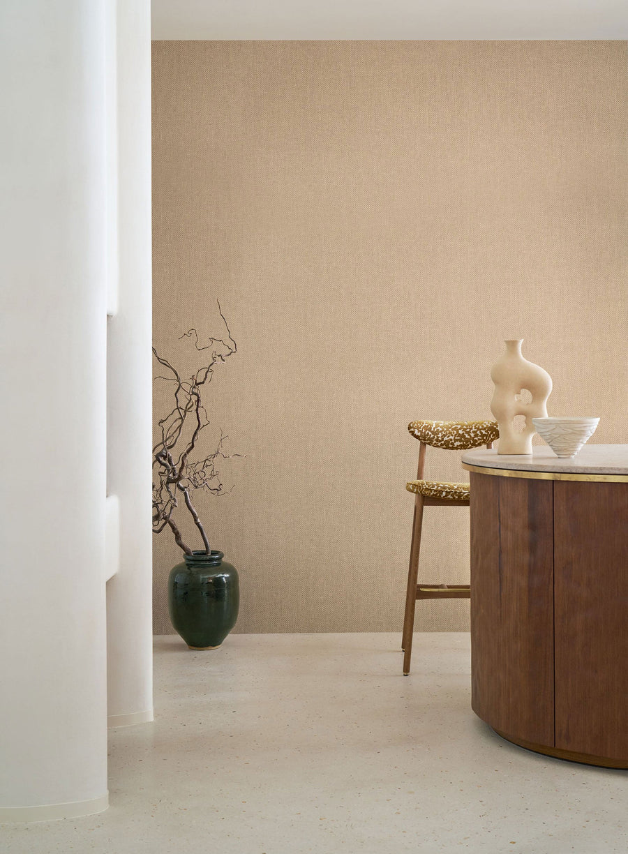 Vannora Sable wallpaper by Casamance, a woven raffia texture in warm sand wrapping a quiet corner with a green vase and a wooden stool