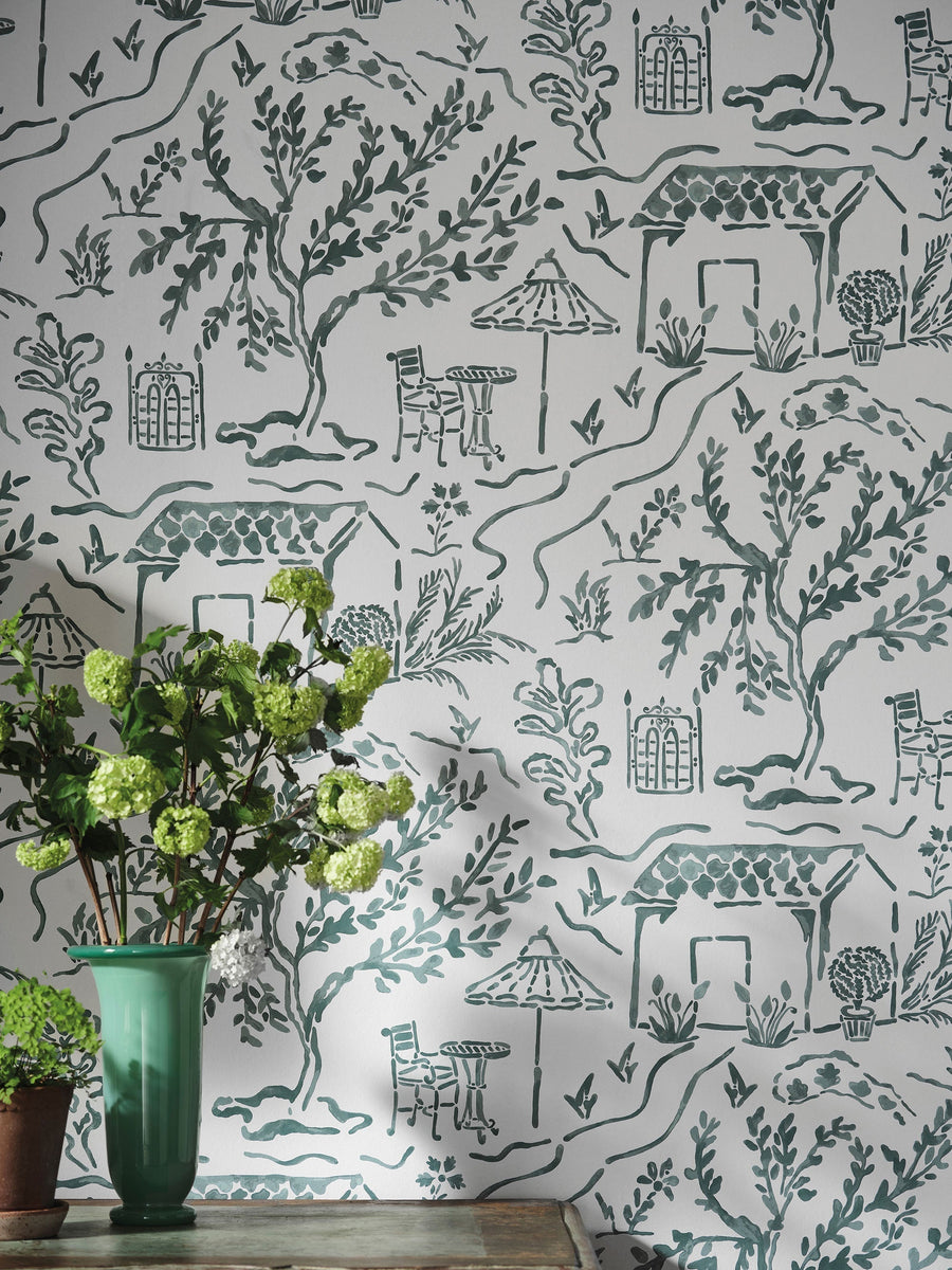 La Quintinie Indigo wallpaper by Nina Campbell, a blue line drawn garden of tiled huts and topiary behind a green vase of hellebores