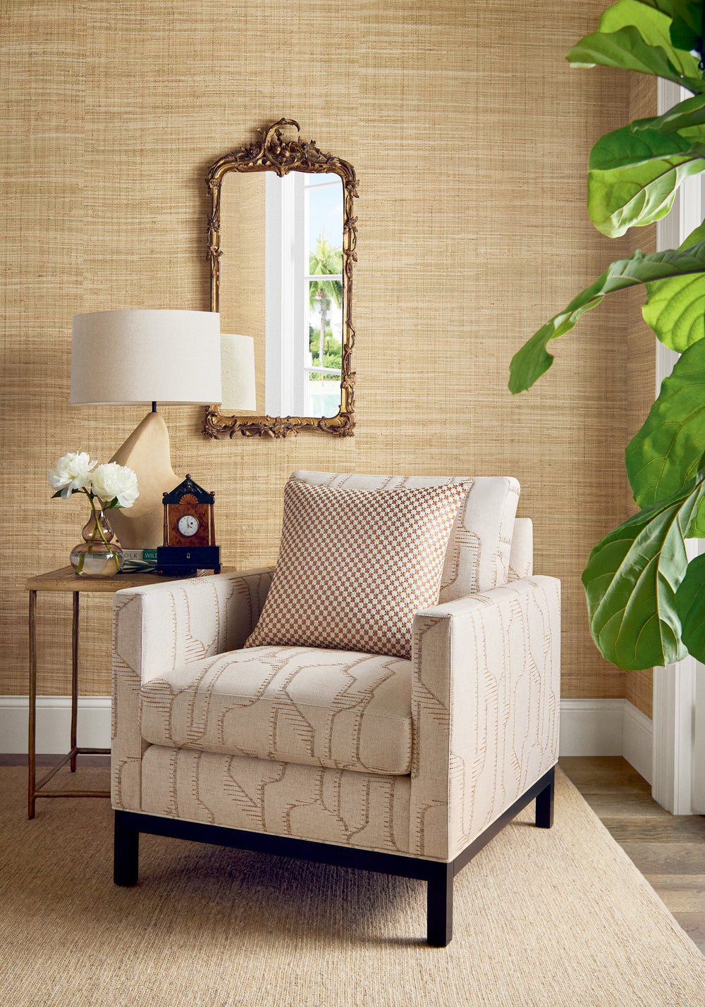 Mackinac natural grasscloth by Thibaut, a honey toned woven grass wall behind a cream patterned armchair, a carved gilt mirror and a fiddle leaf fig