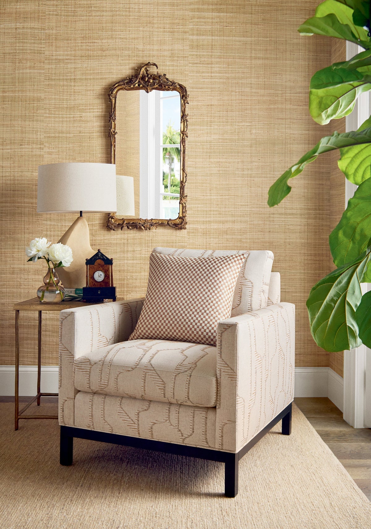 Mackinac natural grasscloth by Thibaut, a honey toned woven grass wall behind a cream patterned armchair, a carved gilt mirror and a fiddle leaf fig