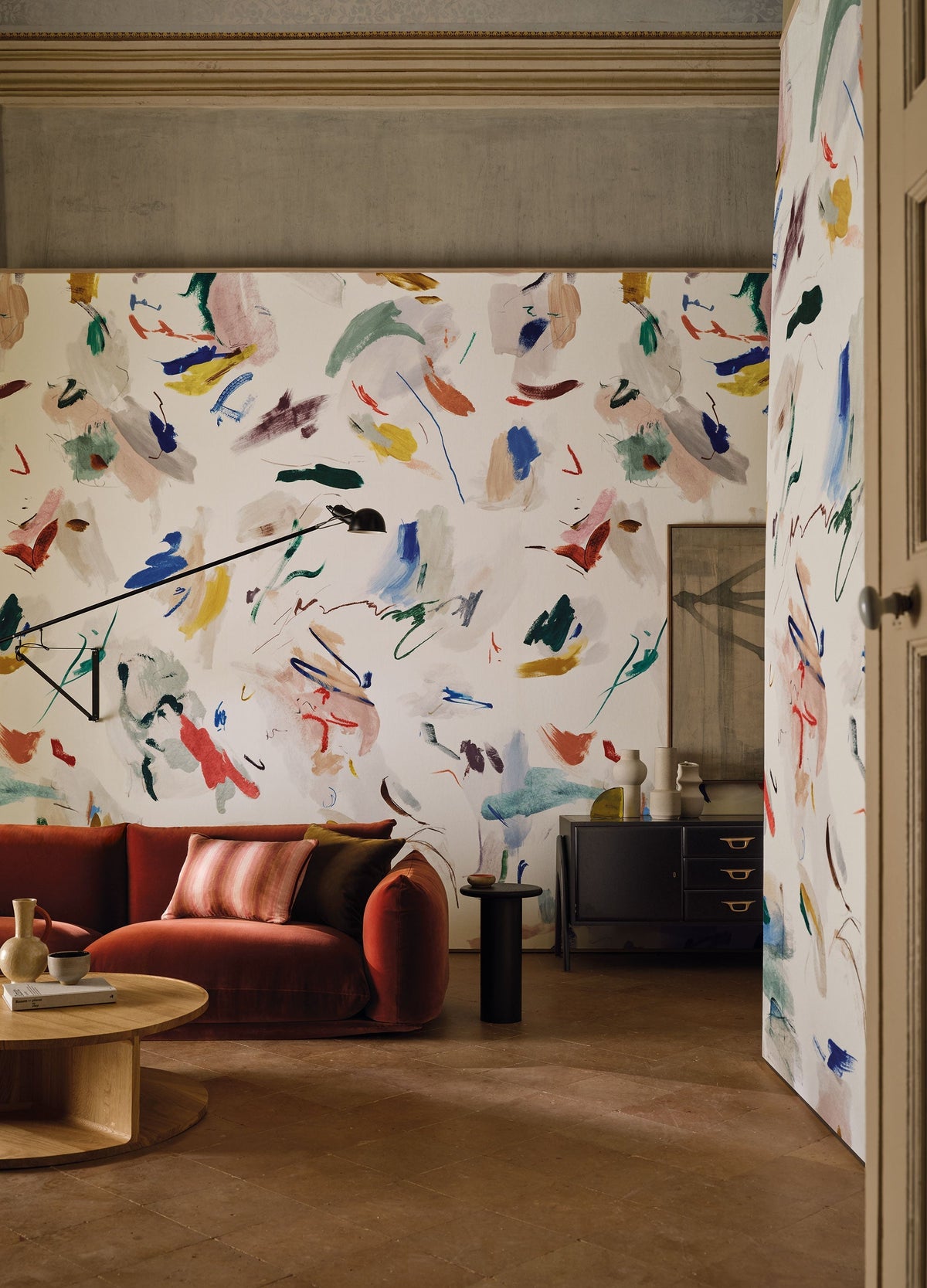 Pigment Multi mural by Black Edition, scattered multicoloured brushstrokes on cream wrapping a living room with a red velvet sofa and an open doorway