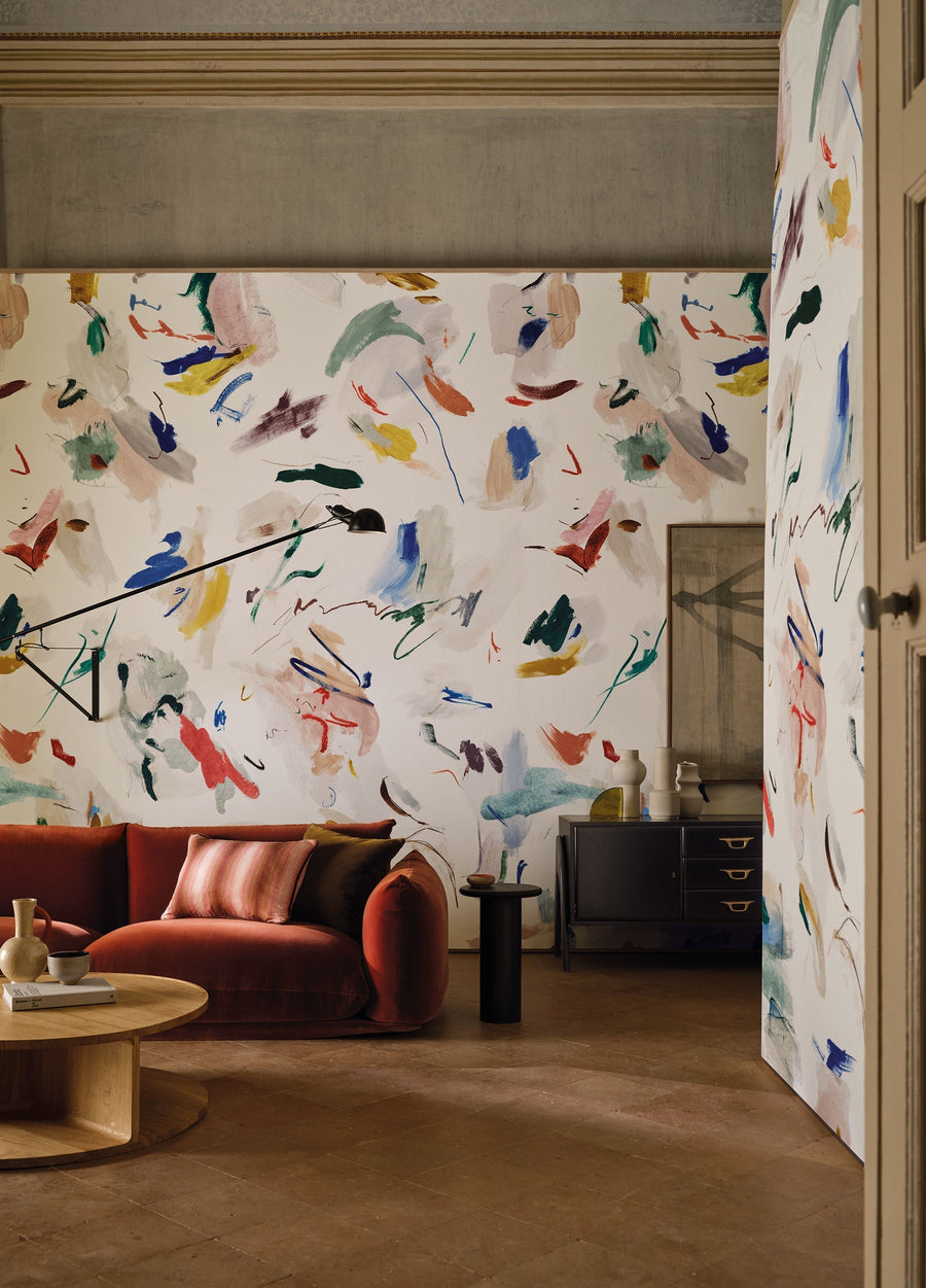Pigment Multi mural by Black Edition, scattered multicoloured brushstrokes on cream wrapping a living room with a red velvet sofa and an open doorway