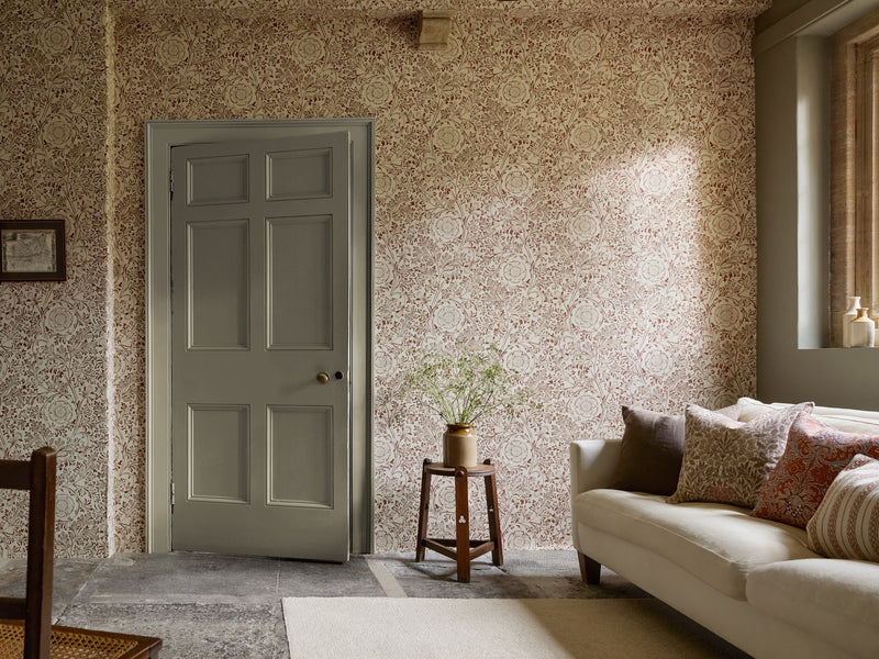 Chamomile in Flax and Madder from The Unfinished Works by Morris & Co, a dense cream and madder floral covering a sitting room with a pale sofa, a sage painted door and a brass plant stand
